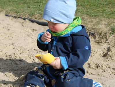 Foto aus der Kinderkrippe: Ein Kleinkind sitzt im Sand mit einem Schäufelchen in der einen Hand und betrachtet Sandkörnchen in der anderen Hand