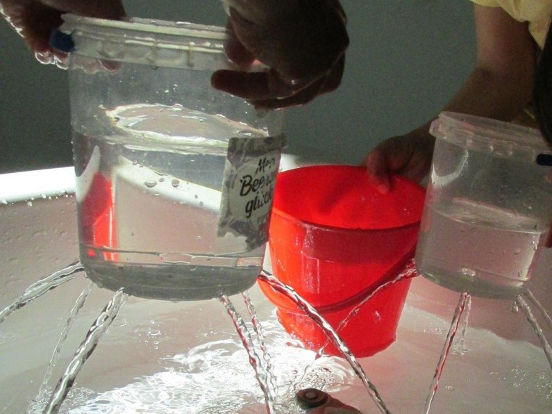 Behälter mit Wasser gefüllt und Löchern aus denen Wasser in ein Becken strömt