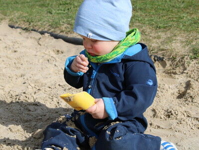 Foto aus der Kinderkrippe: Kleinkind beim Spielen im Sand Foto aus der Kinderkrippe: Ein Kleinkind sitzt im Sand mit einem Schäufelchen in der einen Hand und betrachtet Sandkörnchen in der anderen Hand