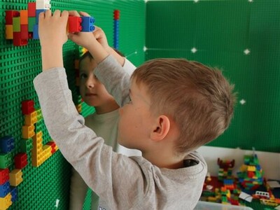 Foto aus dem Kindergarten: Junge spielt Lego an einer Legowand Foto aus dem Kindergarten: Ein Junge steht an einer Wand mit grünen Bauplatten von Lego und setzt Legosteine darauf