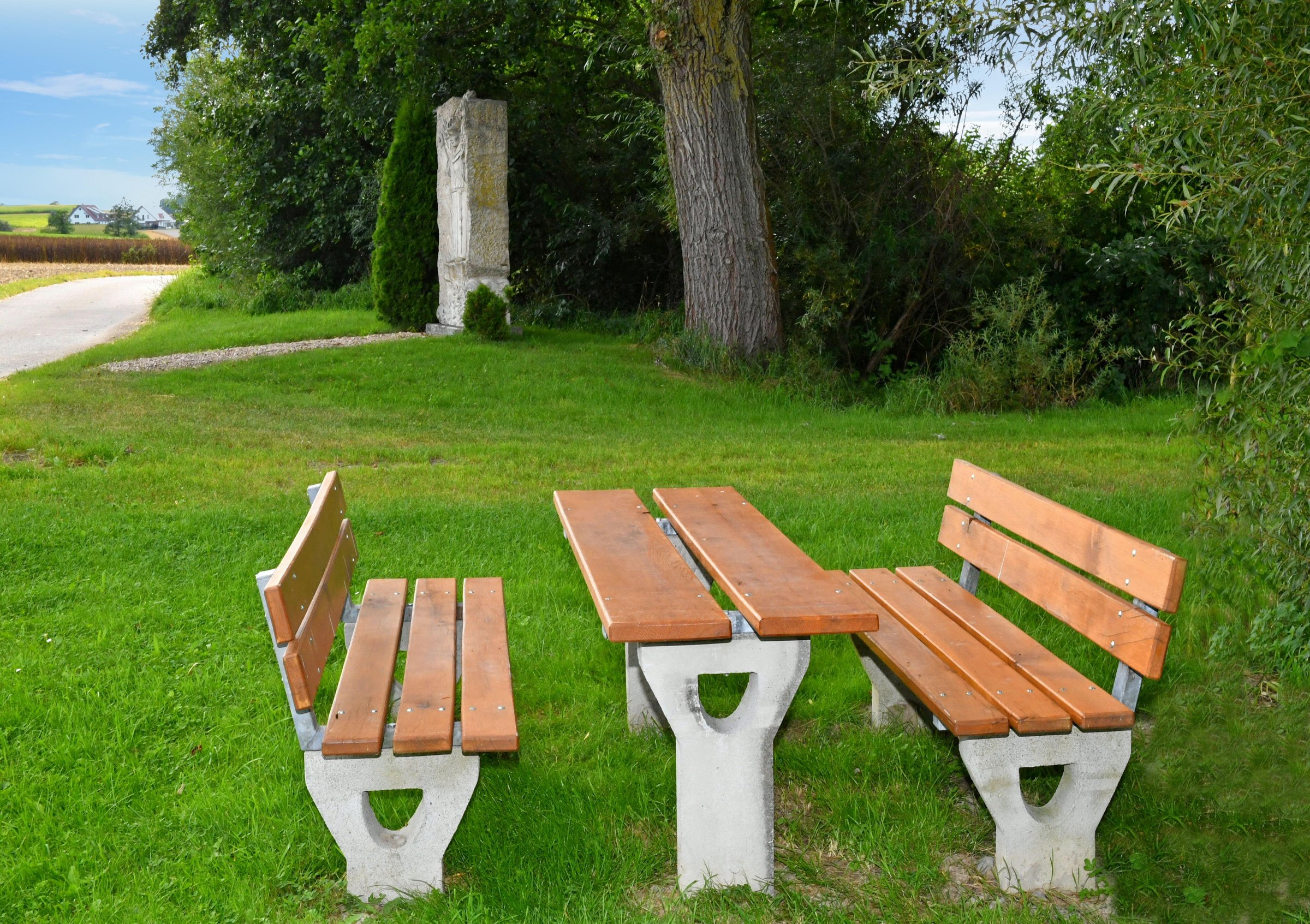 Eine Sitzgelegenheit mit zwei Bänken und einem Tisch in der Mitte. Ringsrum sind Bäume, Büsche, ein Feldweg und eine Skulptur zu sehen.