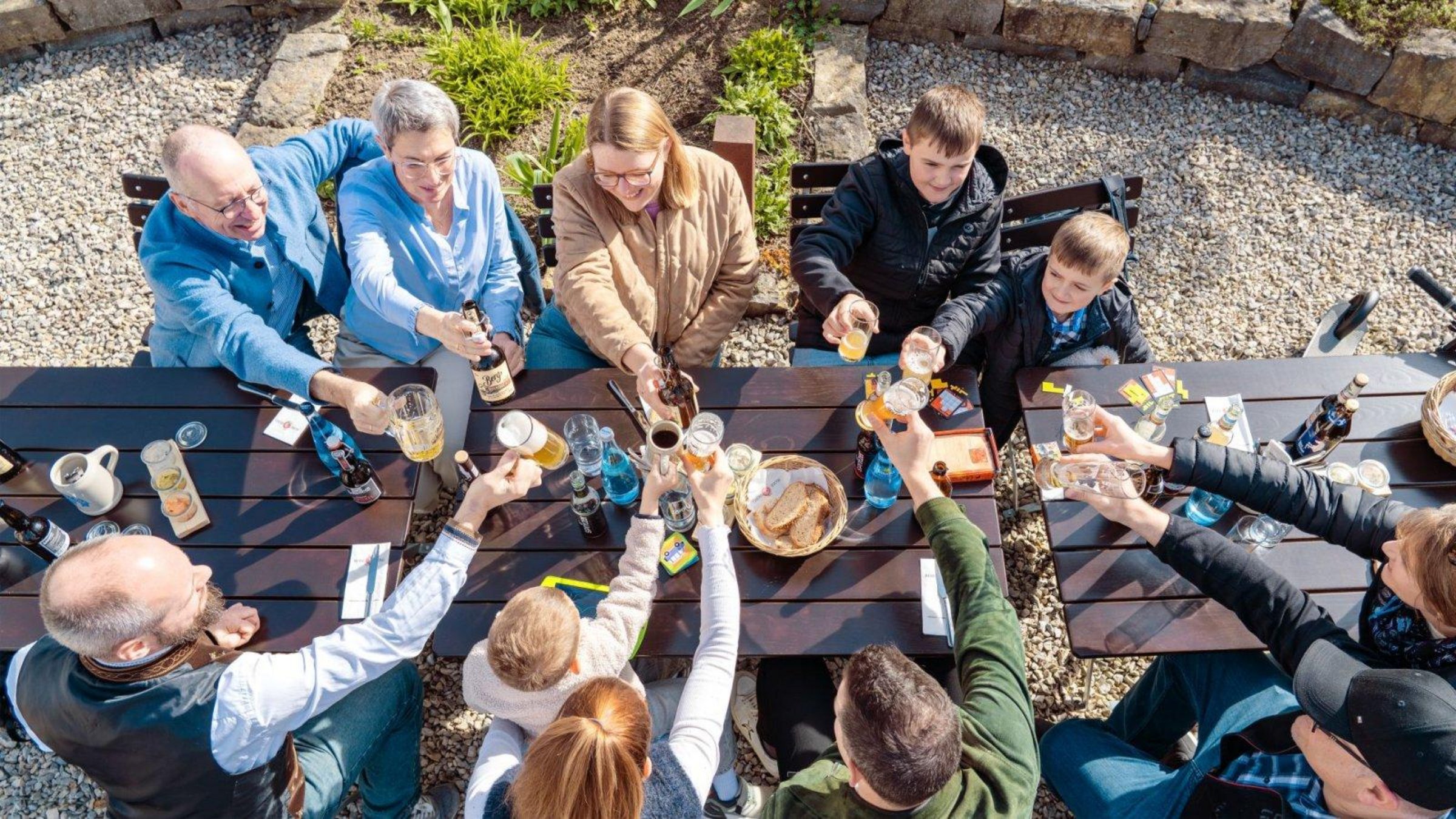 Mehrere Personen jeglichen Alters sitzen im Freien gemeinsam am Tisch und stoßen miteinander an. 
